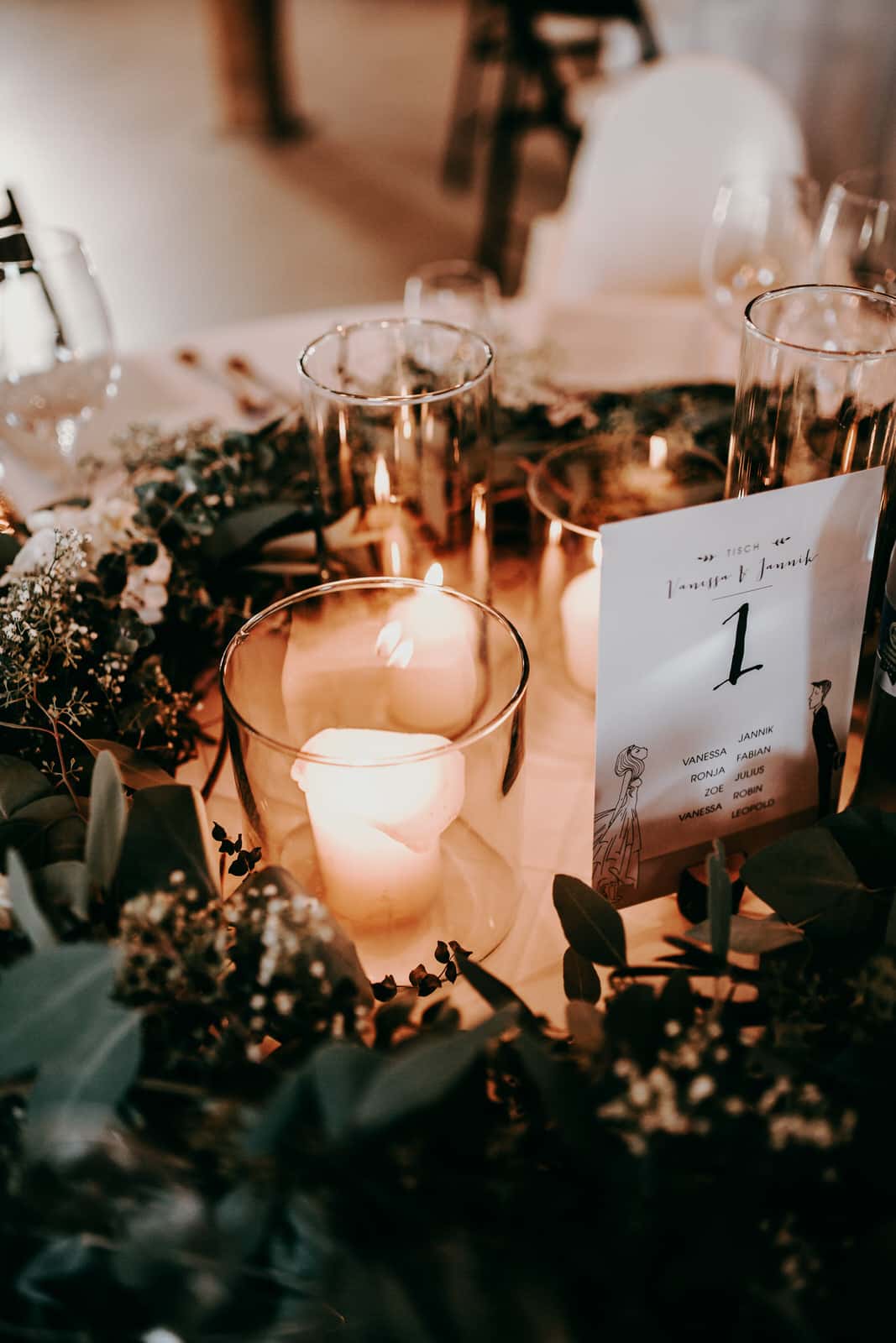 Als Hochzeitsfotografen lieben wir Details.Zu sehen ist ein Ausschnitt die Tischdekoration einer Hochzeitsfeier: weiße Stumpferen in einem edlen Glas, das Kerzenlicht flackert und gibt dem danebenliegenden Eukalyptus einen besonderen Farbton. Die Tischkarte zeigt die Tischnummer an und die Namen der Hochzeitsgäste, welche sich an diesem Tisch einfinden sollen.